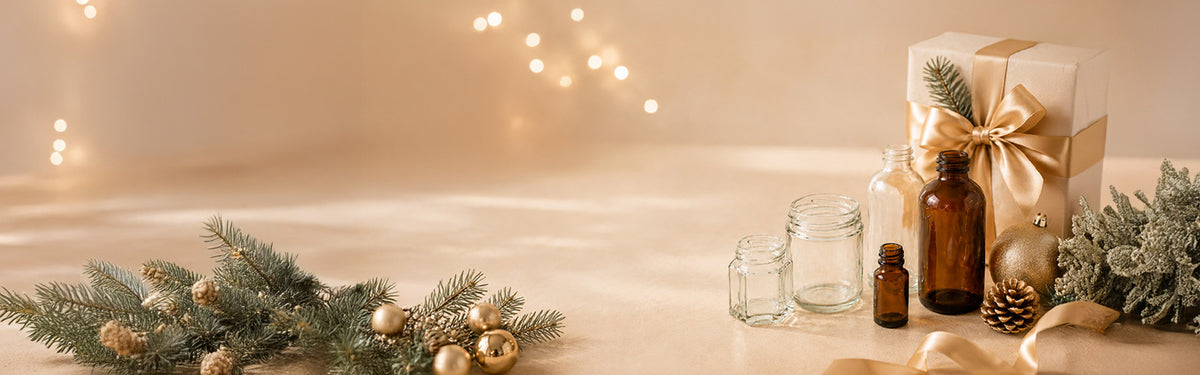 A styled holiday tabletop scene with amber glass bottles, clear glass jars, pine branches with gold ornaments, a wrapped gift box tied with a gold ribbon, and warm string lights in the background.